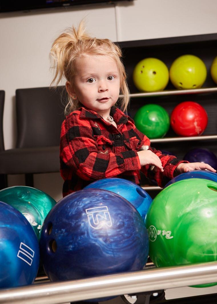 Young child in red plaid shirt standing among colorful bowling balls
